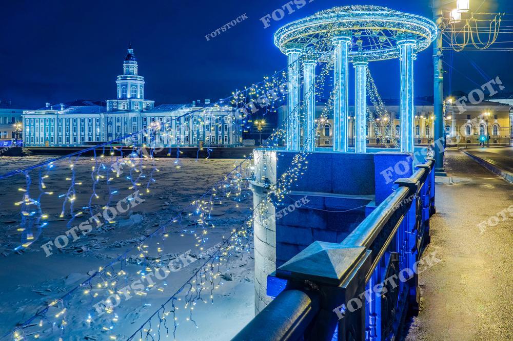 На фотографии изображена набережная реки Невы в Санкт-Петербурге, украшенная праздничными огнями к Новому году. Вдалеке виднеется здание Кунсткамеры, одного из старейших музеев России. Замерзшая поверхность Невы дополняет зимний пейзаж, а огни ночного города отражаются в снегу и льду, создавая яркий и праздничный вид.